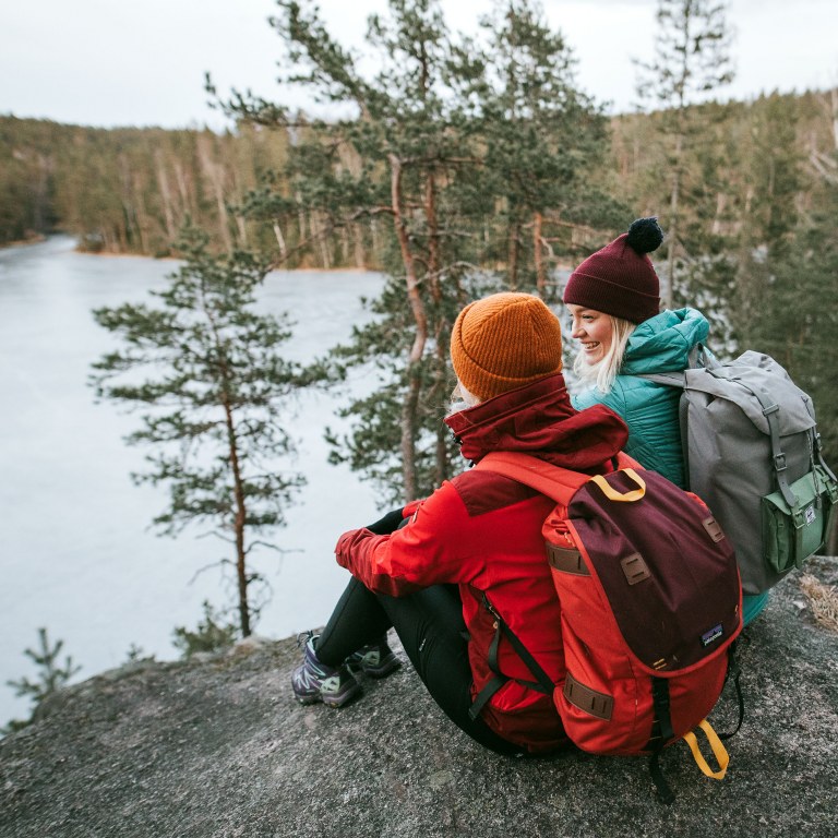 Two beanie-headed people sit on a rock and a forest lake is visible below.