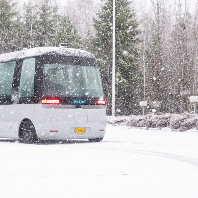 A shuttle bus driving in and leaving tracks in the snow with snow falling down.