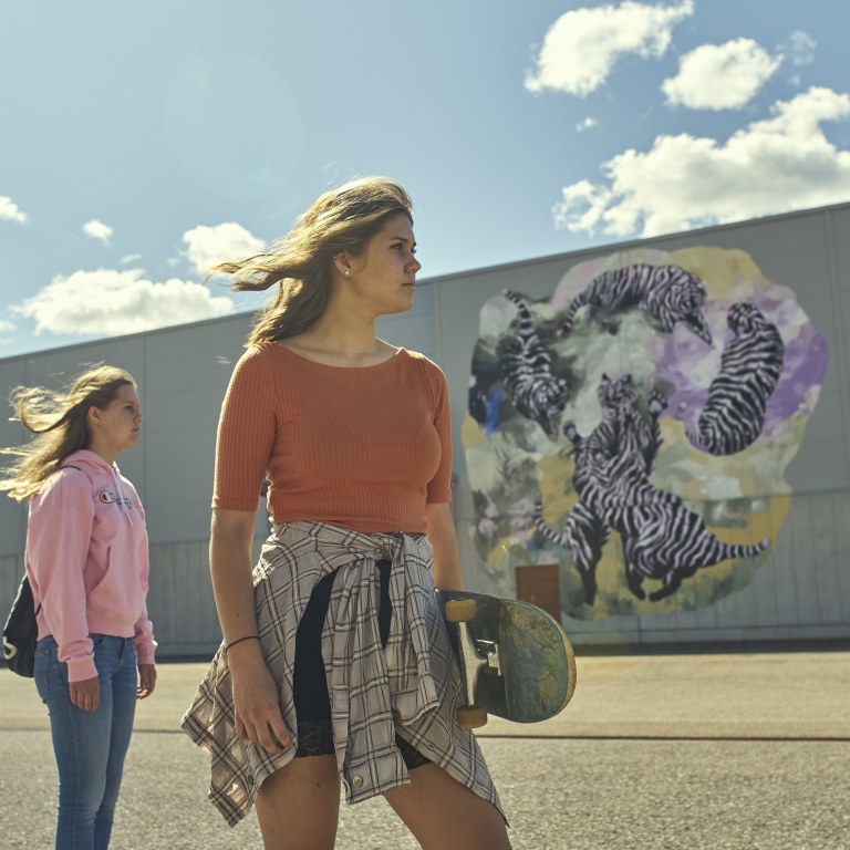 Two girls standing sideways in front of a gray wall with murals.