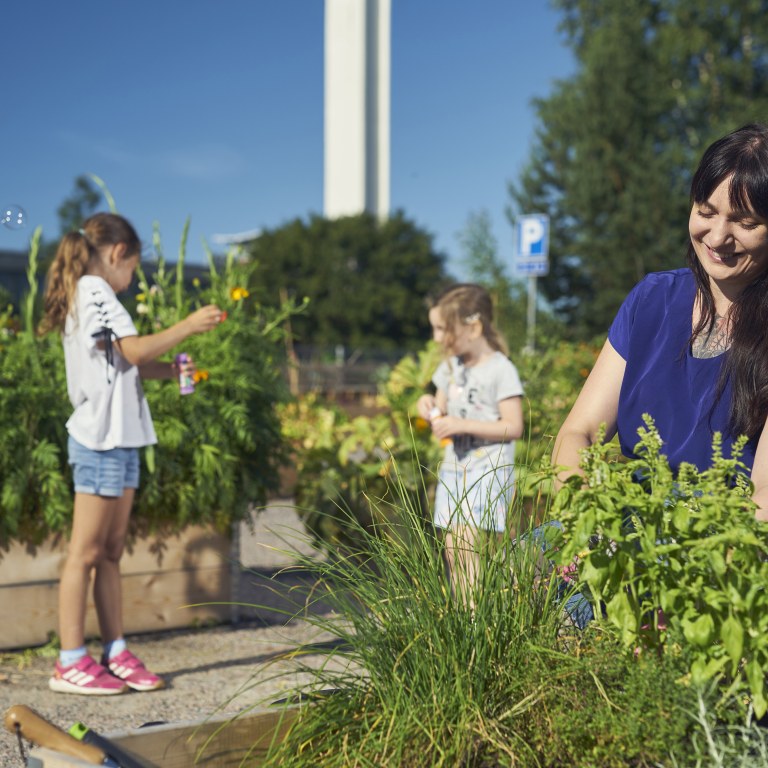 A woman planting herbs and smiling with two girls blowing soap bubbles in the background.