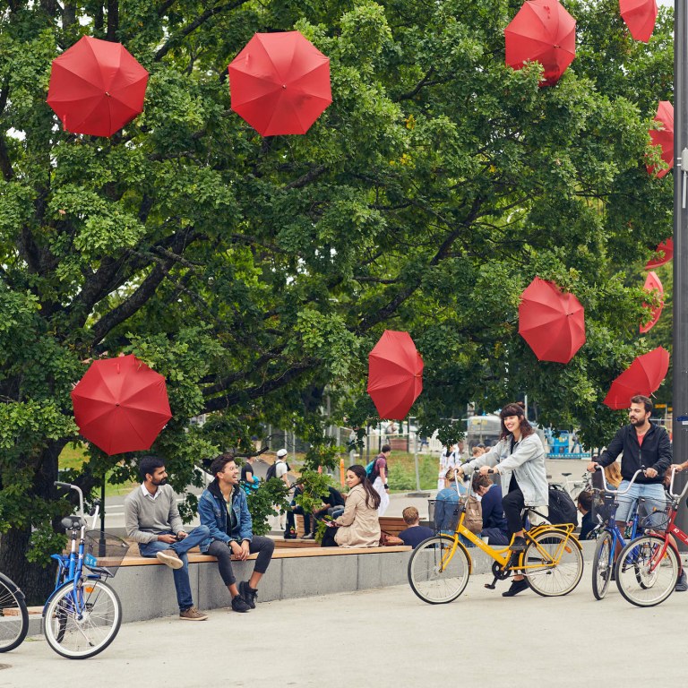 A big green oak tree that has red umbrellas attached to its branches with people sitting and biking. 