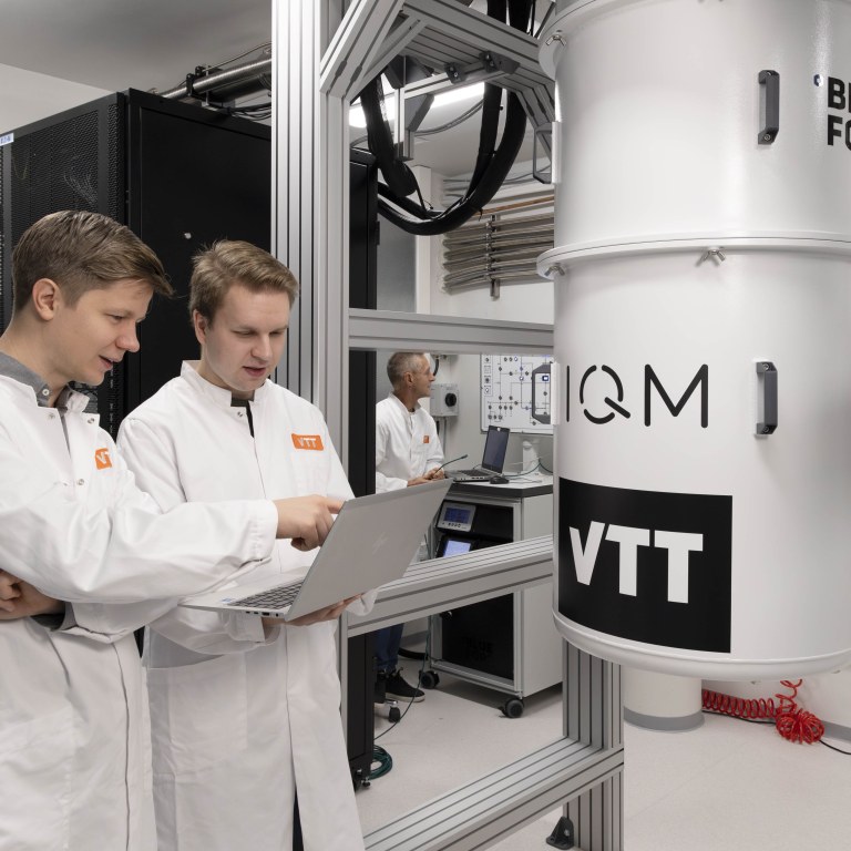 Two scientists with VTT logos on their lab coats looking and pointing at a laptop in a cleanroom next to a quantum computer, with a third scientist in the background.