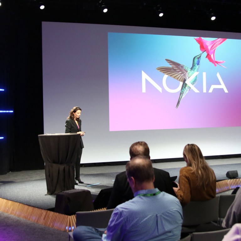 A woman talking on stage in front of a screen that has the image of a humming bird and Nokia written on it. The room is dark but lit with blue tubular lights on the wall and purple spotlights. People are sitting in the audience on the right of the picture.