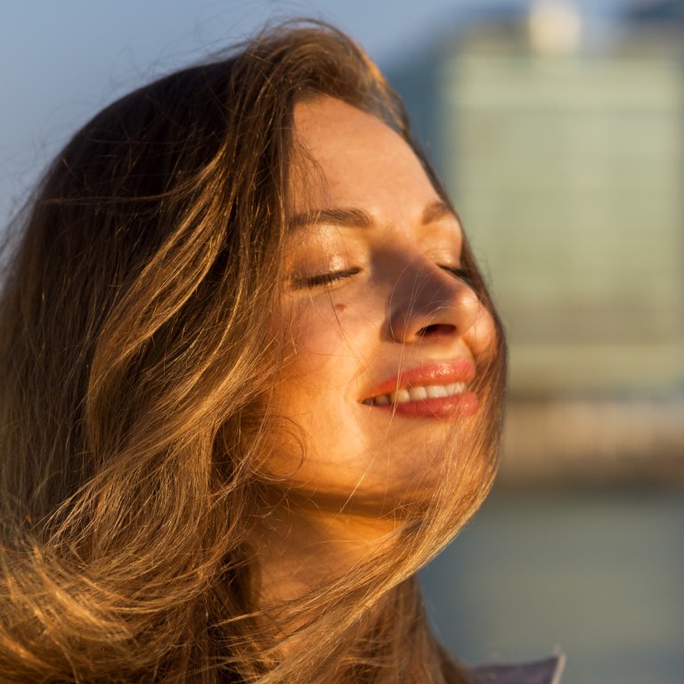 A woman with her eyes closed, her face basking in the sun and her brown hair flowing in the wind.