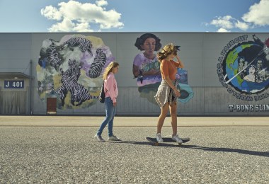 Two girls walking and skateboarding in front of a gray wall with murals. 