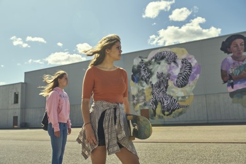 Two girls standing sideways in front of a gray wall with murals.