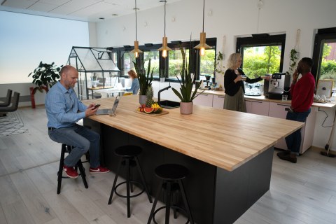 A man sitting at a kitchen island with two people talking and drinking coffee in the on the right side of the island. 