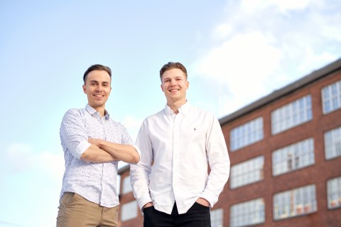 Two men posing and smiling for camera with a red brick building in the background on the right