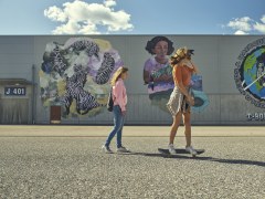 Two girls walking and skateboarding in front of a gray wall with murals. 