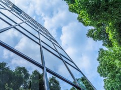 Glass office building pictured skywards with blue clouds and green leaves