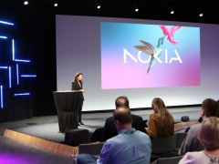 A woman talking on stage in front of a screen that has the image of a humming bird and Nokia written on it. The room is dark but lit with blue tubular lights on the wall and purple spotlights. People are sitting in the audience on the right of the picture.